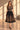 A woman models a black midi dress with gold floral embroidery, featuring a sheer embroidered bodice, short sleeves, a defined waistband, and a full flared skirt, set against a warm beige background with window shadow patterns.